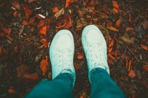 Person wearing white sneakers standing on autumn leaves scattered on the ground.