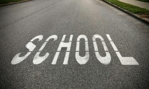 The word 'SCHOOL' painted in large white block letters on an asphalt road near a sidewalk, serving as a traffic warning for a nearby school zone.